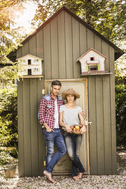 Portrait of couple leaning against shed, young woman holding basket of apples - Foto, Imagen