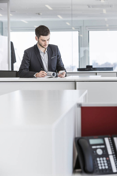 Businessman standing in office reading paperwork and using smartphone - Foto, Bild