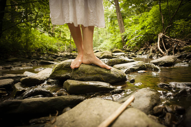 Bare feet of mid adult woman wearing white dress balancing on rock in riverbed - Foto, Imagem