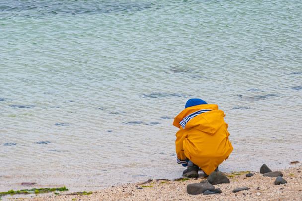 Lapsi, jolla on tuulitakki kyyristyneenä meren rannalla
 - Valokuva, kuva