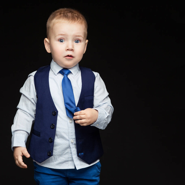Cute little boy in blue business suit with surprised expression on face, touching his tie. Over dark background. - Zdjęcie, obraz