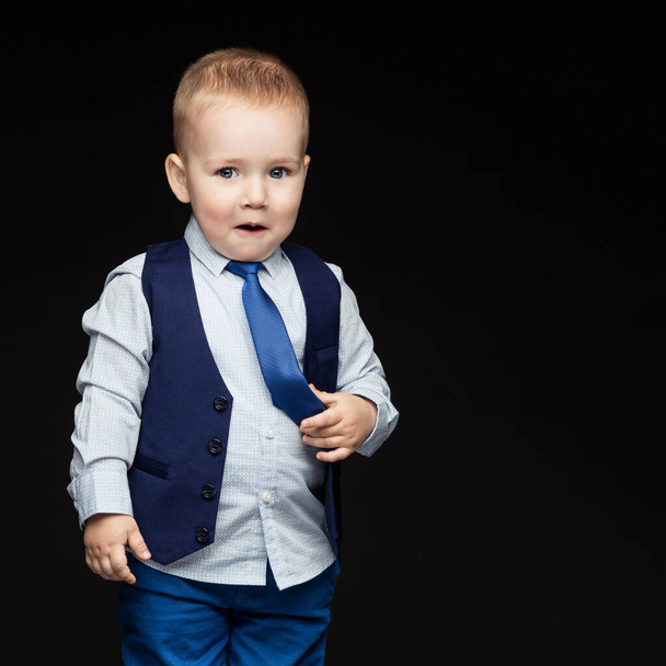 Handsome shy little boy in business suit with emotional expression on face touching his blue tie. Over dark background. Square composition. Copy space. - Foto, Bild