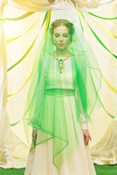 Beautiful young woman in yellow dress covered with green veil. Wedding arch on background. - Foto, Imagen