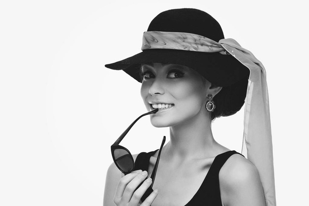 Beautiful young woman looking like Audrey Hepburn in hat with scarf and holding sunglasses - 写真・画像