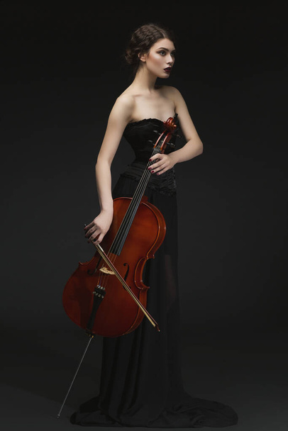 Beautiful young woman standing in black long skirt and corset with cello. Over black background. - Фото, зображення