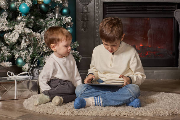 Small children with computer tablet are sitting near Christmas tree at home. Merry Christmas and Happy New Year. - Foto, Imagen