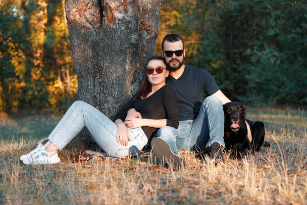 Young beautiful couple of their Labrador are sitting on the grass in the Park. Warm autumn evening in the forest. The concept of a happy family life in love. - 写真・画像