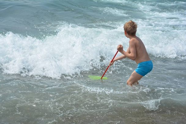 little blond boy with blue swimsuit stands with a net in the water and wait for the sea - wave\n - Photo, Image