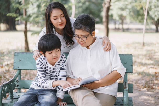 Happy Asian family with mother, grandmother and son sitting on  the bench while read book in the park - Фото, изображение