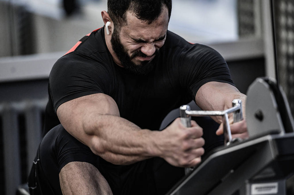 strong young bearded caucasian male in black jersey wearing wireless headset lifting heavy weight in sport gym with grimace of stress pain on his face - Фото, зображення