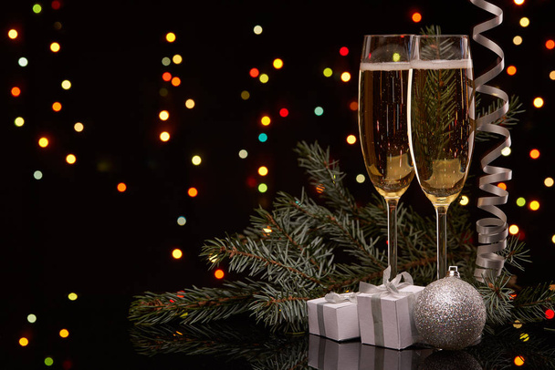 Two glasses with champange, fir tree branch with decoration, gift boxes on a black background with multi-colored lightes of garland.  New year and Christmas. - Zdjęcie, obraz