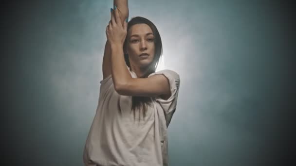 Elastyczna kobieta w białej koszulce tańcząca rękami w dymiącym studio - Materiał filmowy, wideo