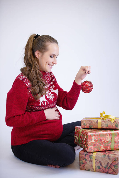 A pregnant girl is sitting on the floor with her hand in her stomach. With his second hand holds a Christmas ball. Nearby are gift boxes. White background - Φωτογραφία, εικόνα