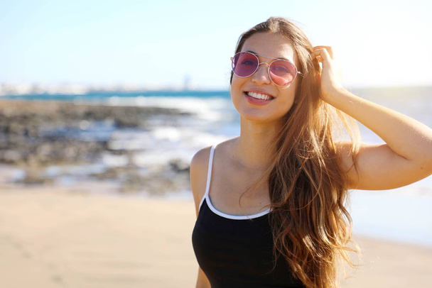 ziemlich moderne, sportliche Frau mit Sonnenbrille, die glücklich lächelnd in die Kamera am Strand blickt - Foto, Bild