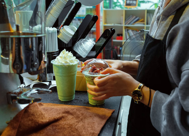 Barista egy kávézóban dolgozik. A kezek becsukják a fedelet. A lányok épp most készítettek hideg matcha teát. Zöld matcha és fehér krém - Fotó, kép