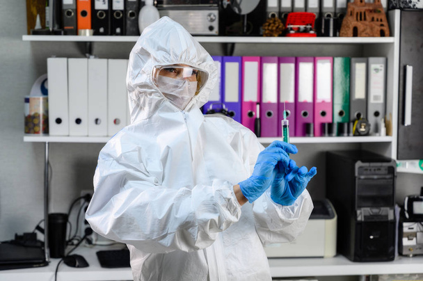 Photo of a doctor in a white medical protective suit in an office. He stands in front of the camera with a syringe in his hands, preparing to give an injection to the patient. - Photo, Image