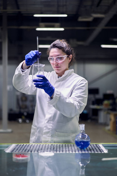 Serious lab girl in glasses and white coat with experimental glass in her hands conducts experiments - Фото, изображение