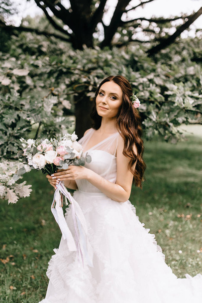 Beautiful bride with bouquet flowers stands on forest background.  beautiful bride in delicate dress outdoors. Close up portrait of young bride in park in sunny weather wooded area. - Foto, Imagem