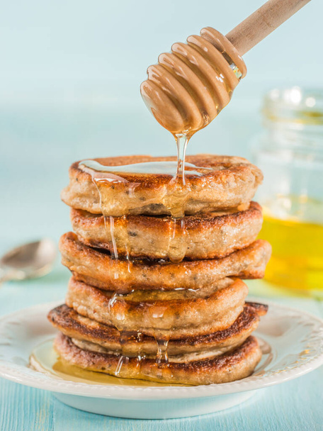 Stack of small pancakes with banana and chia seeds in honey on light blue background. Honey pouring over stack of mini pancakes with chia or poppy seeds. - Foto, immagini