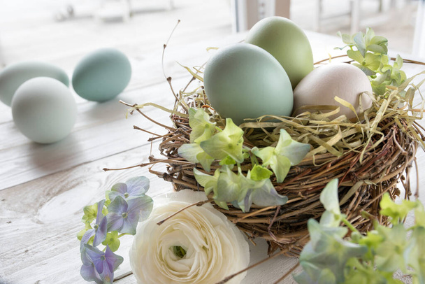 Still life with pastel colored easter eggs in a nest of straw and flowers on a white wooden table - Foto, Bild