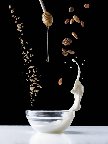 Falling granola with milk splash, falling nuts and honey flows from dipper. Healthy breakfast ingredients. Flying food - Photo, image