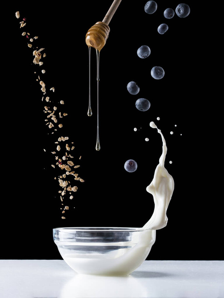 Falling granola with milk splash, falling blueberries and honey flows from dipper. Healthy breakfast ingredients. Flying food - Фото, изображение
