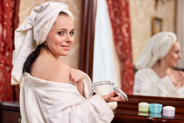 Young woman with cosmetic cream sits before a house  dressing table..Concept body care. - Zdjęcie, obraz