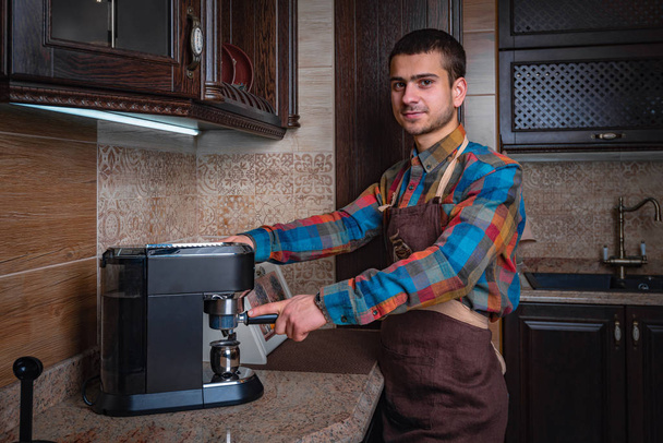 Стильный молодой человек с бородой, одетый в повседневную одежду, готовит кофе в кофеварке
. - Фото, изображение