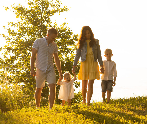 海の近くを歩く幸せな家族。田舎の野原と木々。日没や日の出の暖かい色。愛する両親と美しい子供たち。愛と親子の概念. - 写真・画像