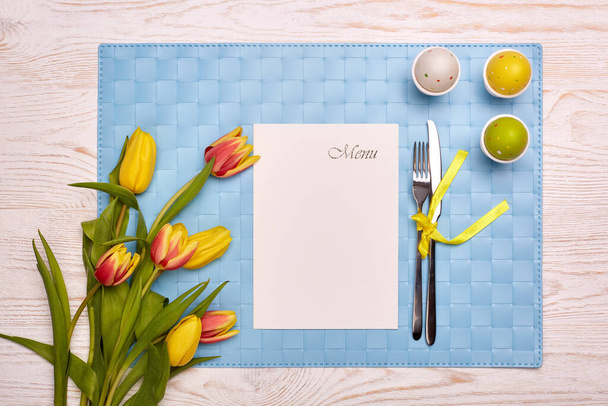 Easter table setting with spring tulips flowers, colorful egg, tableware and blue napkin on a wooden table. Top view. - Photo, image