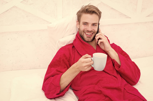 Small talk over his favorite drink. Happy man talking on phone with cup of hot drink in bed. Bearded handsome guy enjoying his drink break in bedroom. Starting his day with tasty drink - Photo, Image