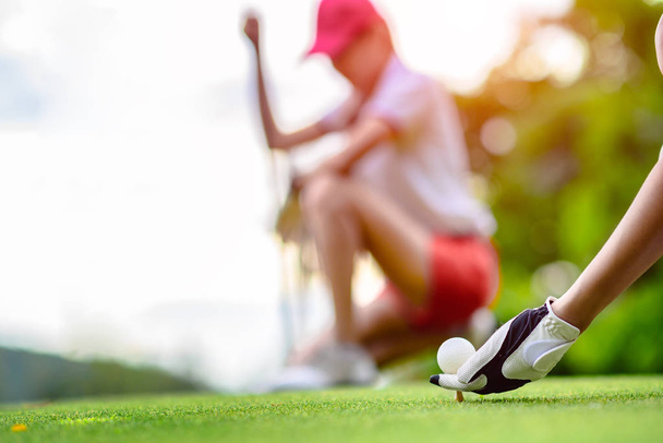 hand of young woman golf player holding golf ball laying on wooden tee, prepare and ready to hit the ball to the destination target, opponent competitor or golf mate buddy watching in background - Foto, afbeelding