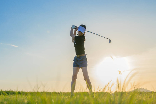 mujer jugador de golf en acción se está configurando para golpear la pelota de golf lejos de fairway al destino verde apagado, fairway a la luz del atardecer
 - Foto, Imagen