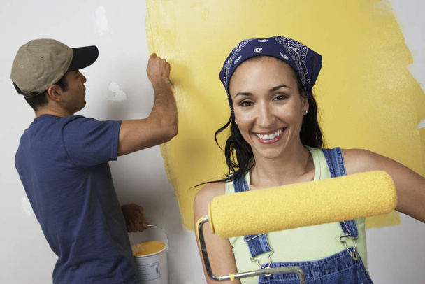 Portrait of happy young woman holding roller paint with man painting wall in background - Foto, Bild