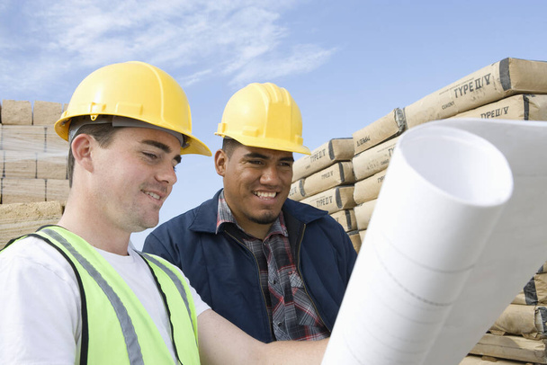 Two workers with blueprint standing in front of cement bags and bricks - Photo, image