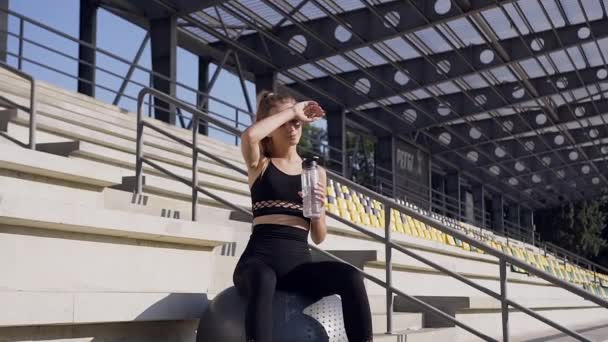Pretty tired athletic young woman in fitness clothes sitting on big gymnastic ball and drinking water during sport workout outdoors - Metraje, vídeo