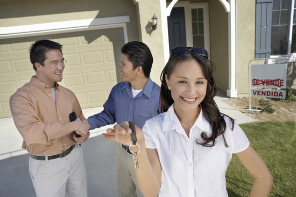 Portrait of young woman holding keys while man shaking hands with agent in background - Foto, Imagem
