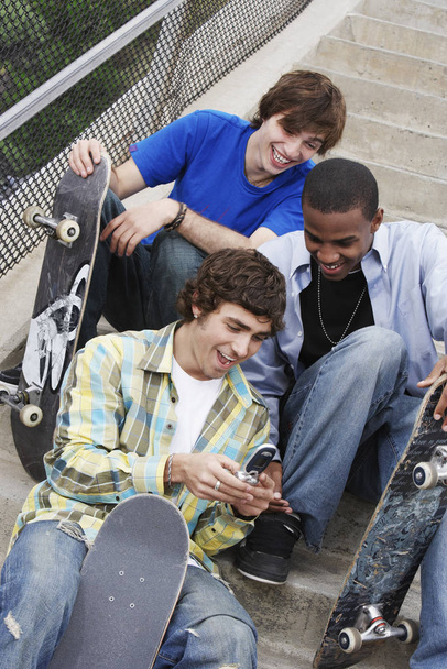 Three young male friends with skateboards reading text message through cell phone - Foto, Imagem