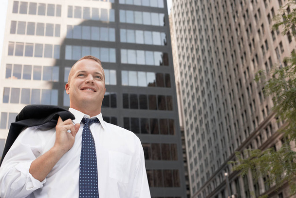 Relaxed mature businessman with coat over shoulder in front of city buildings - Photo, Image