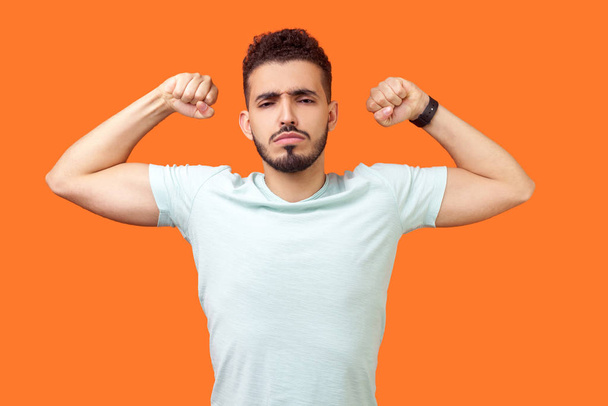 I am strong and powerful. Portrait of confident successful brunette man with beard in white t-shirt showing biceps, full of motivation and willpower. indoor studio shot isolated on orange background - Фото, зображення