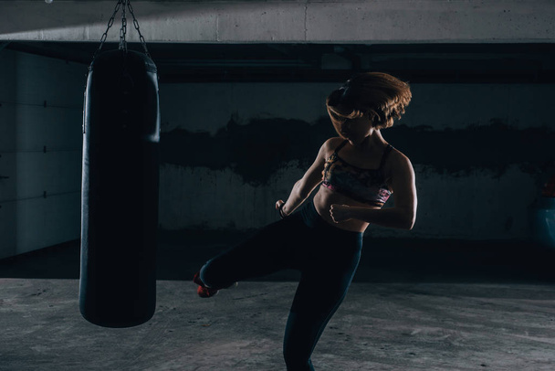 Fiatal sziluett női gyakorló magas rúgás a boxzsák a garázsban. - Fotó, kép