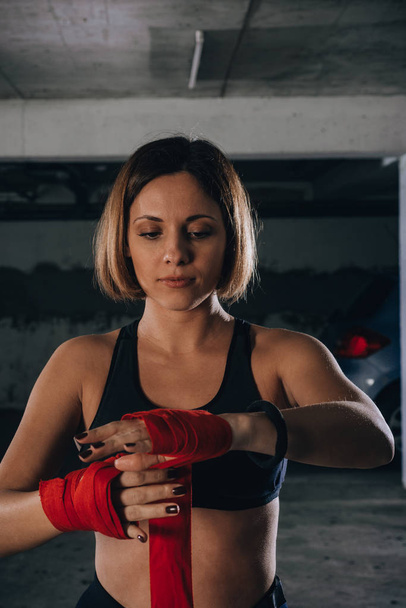 Retrato de close-up de uma mulher bonita fazendo bandagens de boxe vermelho em uma garagem
. - Foto, Imagem