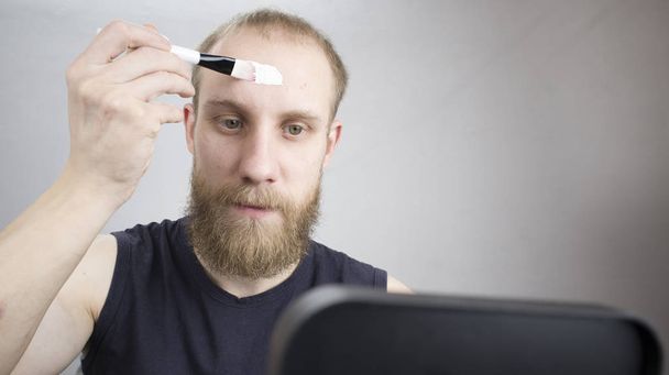 un homme avec une barbe est assis devant un miroir à la maison et met un masque sur son visage pour nettoyer la peau
 - Photo, image