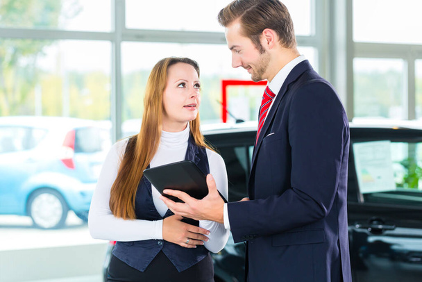 Seller or car salesman and female client or customer in car dealership presenting the interior decoration of new and used cars in the showroom on tablet computer - Foto, Imagen