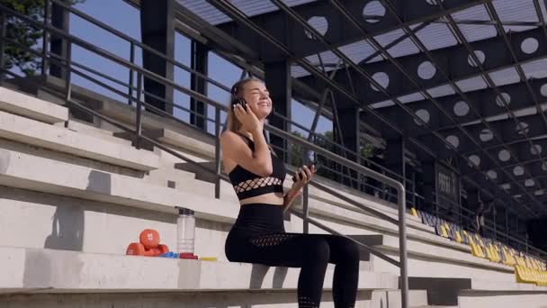 Attractive smiling sporty young woman in sportswear sitting on the tribune and listening music in earphones after fitness training - Séquence, vidéo