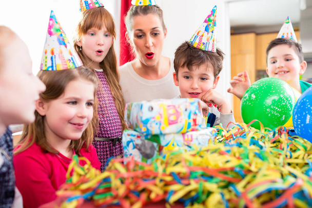 Child unwrapping birthday gift with friends at home birthday party, mom is helping - Photo, image