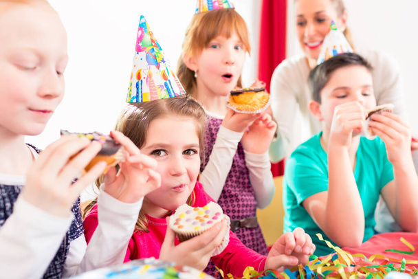 Children grabbing muffins at birthday party and cake, the kids are wearing hats, balloons and paper streamers for decoration - Foto, Imagem