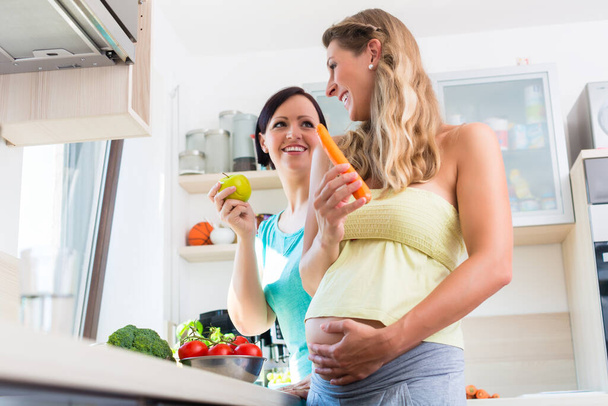 Pregnant woman and girlfriend cooking healthy in domestic kitchen eating a carrot - Fotó, kép