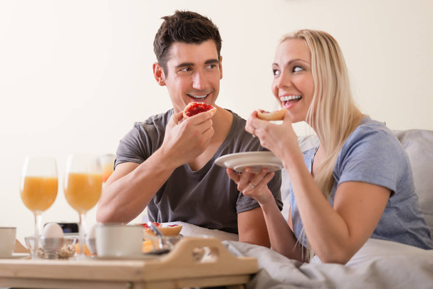 Happy playful couple enjoying breakfast in bed grinning at each other as they prepare to bite into toast and jam with a spread of orange juice, coffee and eggs on a tray in front of them - 写真・画像