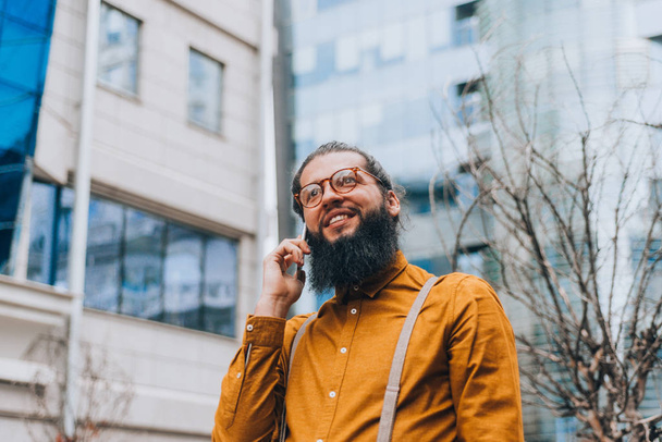 Moderno ragazzo hipster con i capelli lunghi e la barba al lungomare della città avendo conversazione mobile
 - Foto, immagini